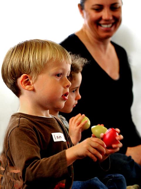 A parent smiles as two young children use egg shakers in a music class for toddlers.