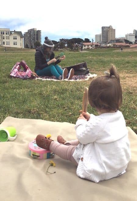 Teacher Rachel tunes a ukulele in the park while a toddler in the foreground holds a rhythm stick.
