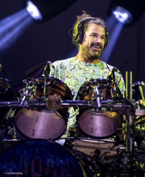 Photo of Jay Lane smiling and playing drums with headphones on