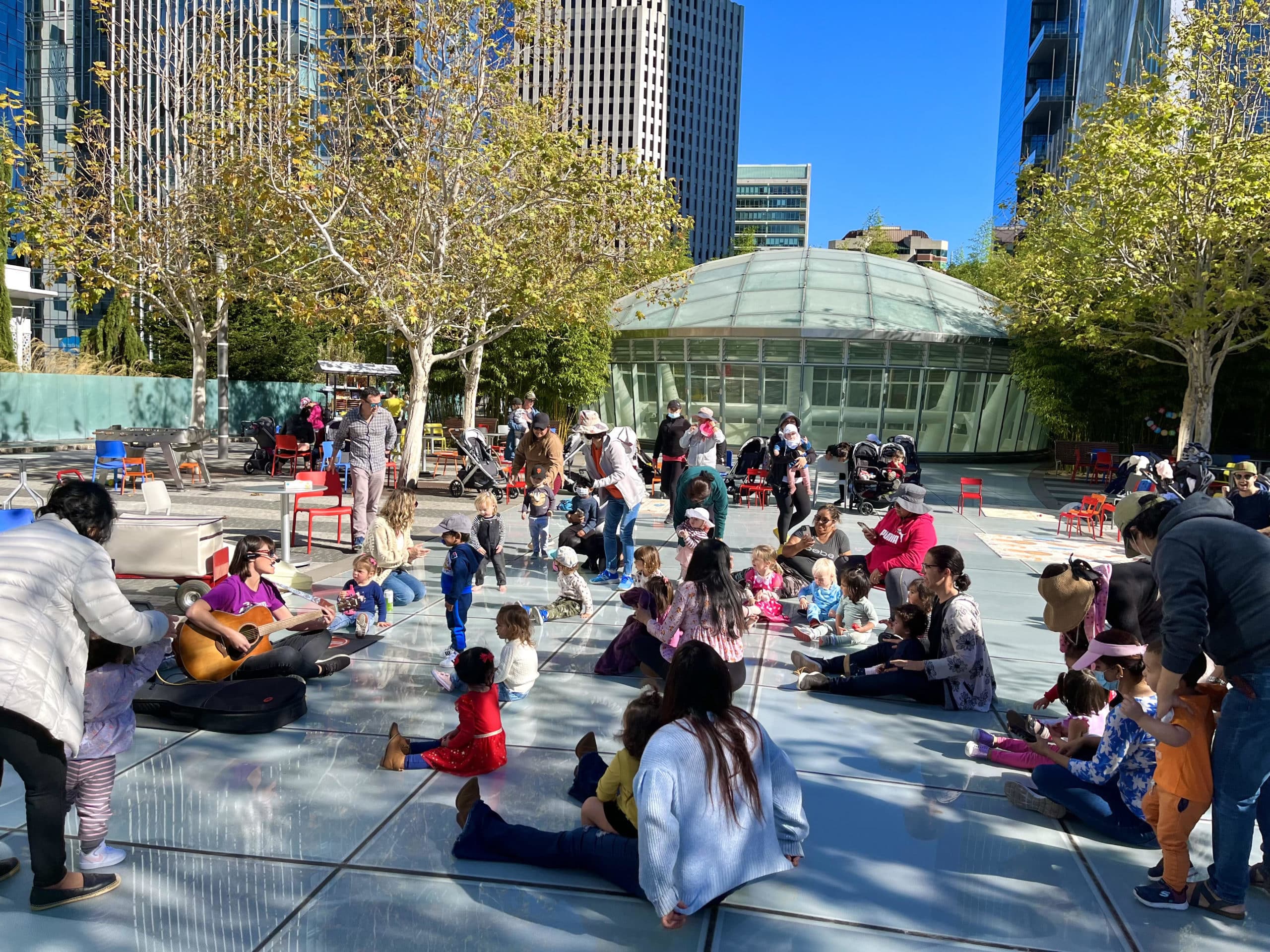 A teacher plays a guitar surrounded by toddlers and parents in Salesforce Park